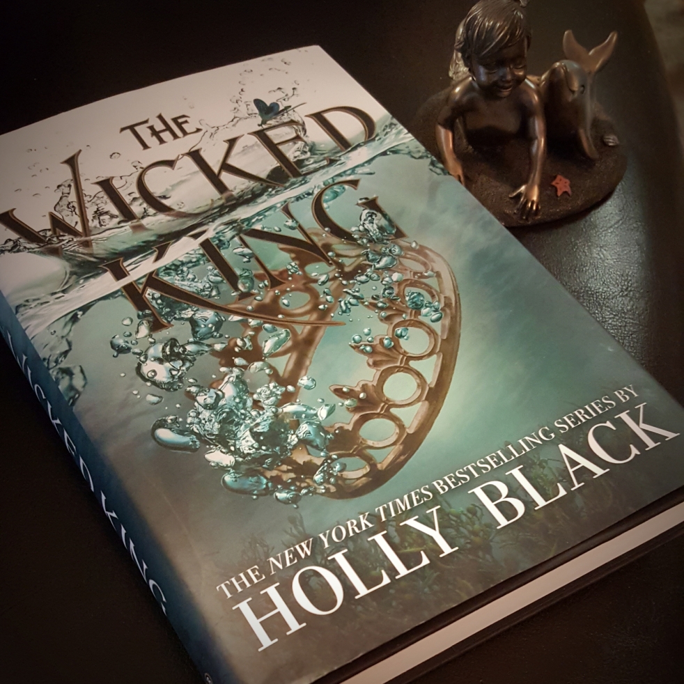 The Wicked King by Holly Black, statuette of boy merman and dolphin beside it